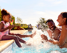 Two young children sit on the edge of a backyard pool enjoying popsicles. They use their feet to splash water at their parents.